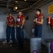 Three military personnel wearing red life vests are standing in what appears to be a naval vessel's interior deck area. Two of the individuals are holding rope lines, while a third appears to be observing or receiving instruction. Safety equipment including a life ring labeled 'Abraham Lincoln' and fire suppression equipment are mounted on the gray bulkhead wall.