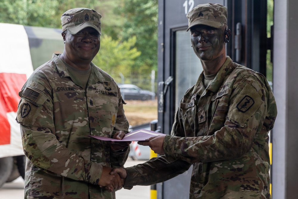 Two U.S. Army soldiers are shaking hands while exchanging what appears to be a document or certificate, likely during an award or recognition ceremony. The soldier on the left, identified by name tape as 'Collins,' wears senior NCO rank insignia, while the soldier on the right has tactical face paint applied. The setting appears to be an outdoor military installation or training area.
