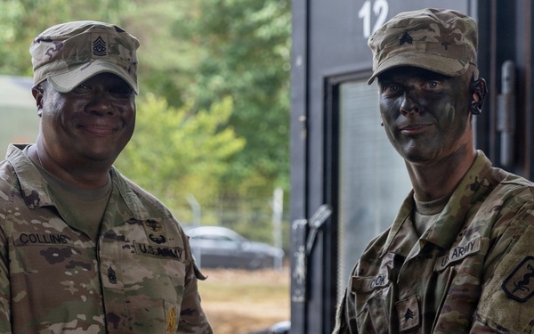 Two U.S. Army soldiers are shaking hands while exchanging what appears to be a document or certificate, likely during an award or recognition ceremony. The soldier on the left, identified by name tape as 'Collins,' wears senior NCO rank insignia, while the soldier on the right has tactical face paint applied. The setting appears to be an outdoor military installation or training area.