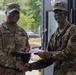 Two U.S. Army soldiers are shaking hands while exchanging what appears to be a document or certificate, likely during an award or recognition ceremony. The soldier on the left, identified by name tape as 'Collins,' wears senior NCO rank insignia, while the soldier on the right has tactical face paint applied. The setting appears to be an outdoor military installation or training area.