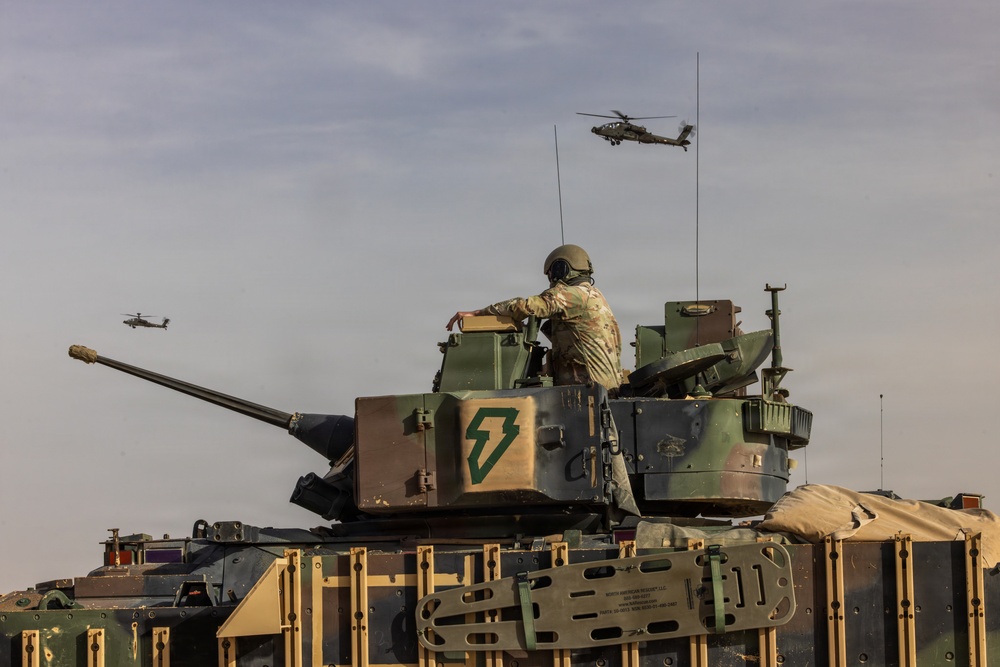 A military scene showing a soldier standing in the hatch of an armored fighting vehicle, likely an M1 Abrams tank or Bradley variant, marked with a green lightning bolt insignia. Two attack helicopters, identifiable as AH-64 Apache variants, are visible flying in the background sky. The scene appears to be a military exercise or operational deployment.