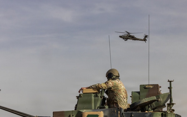 A military scene showing a soldier standing in the hatch of an armored fighting vehicle, likely an M1 Abrams tank or Bradley variant, marked with a green lightning bolt insignia. Two attack helicopters, identifiable as AH-64 Apache variants, are visible flying in the background sky. The scene appears to be a military exercise or operational deployment.
