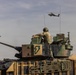 A military scene showing a soldier standing in the hatch of an armored fighting vehicle, likely an M1 Abrams tank or Bradley variant, marked with a green lightning bolt insignia. Two attack helicopters, identifiable as AH-64 Apache variants, are visible flying in the background sky. The scene appears to be a military exercise or operational deployment.