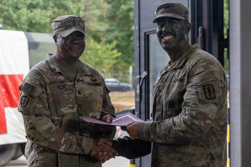 Two U.S. Army soldiers in OCP (Operational Camouflage Pattern) uniforms are shaking hands while jointly holding what appears to be a certificate or award folder. The soldier on the right has tactical face paint applied, suggesting a field exercise or training environment. This appears to be a formal recognition or award ceremony conducted in an outdoor/semi-outdoor military setting.