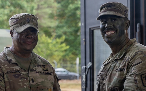 Two U.S. Army soldiers in OCP (Operational Camouflage Pattern) uniforms are shaking hands while jointly holding what appears to be a certificate or award folder. The soldier on the right has tactical face paint applied, suggesting a field exercise or training environment. This appears to be a formal recognition or award ceremony conducted in an outdoor/semi-outdoor military setting.