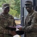 Two U.S. Army soldiers in OCP (Operational Camouflage Pattern) uniforms are shaking hands while jointly holding what appears to be a certificate or award folder. The soldier on the right has tactical face paint applied, suggesting a field exercise or training environment. This appears to be a formal recognition or award ceremony conducted in an outdoor/semi-outdoor military setting.