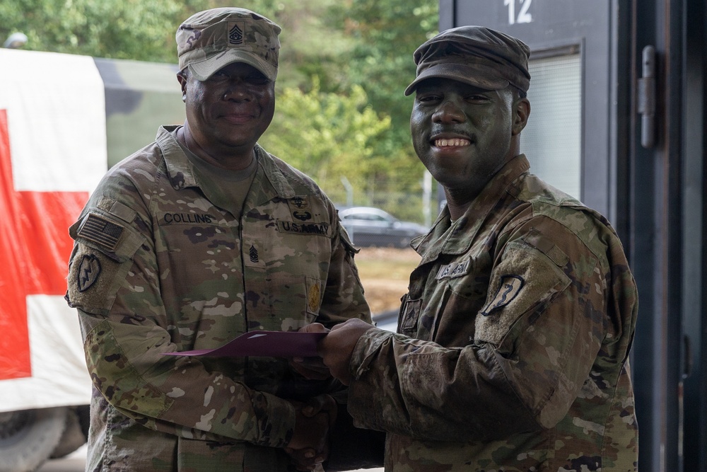 Two U.S. Army soldiers in MultiCam/OCP camouflage uniforms are posing together while one presents a document (likely a certificate or award) to the other. The soldier on the right has camouflage face paint applied, suggesting recent field training activity. The ceremony appears to be taking place at a military installation or training area.