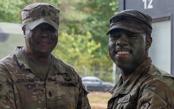 Two U.S. Army soldiers in MultiCam/OCP camouflage uniforms are posing together while one presents a document (likely a certificate or award) to the other. The soldier on the right has camouflage face paint applied, suggesting recent field training activity. The ceremony appears to be taking place at a military installation or training area.