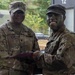 Two U.S. Army soldiers in MultiCam/OCP camouflage uniforms are posing together while one presents a document (likely a certificate or award) to the other. The soldier on the right has camouflage face paint applied, suggesting recent field training activity. The ceremony appears to be taking place at a military installation or training area.