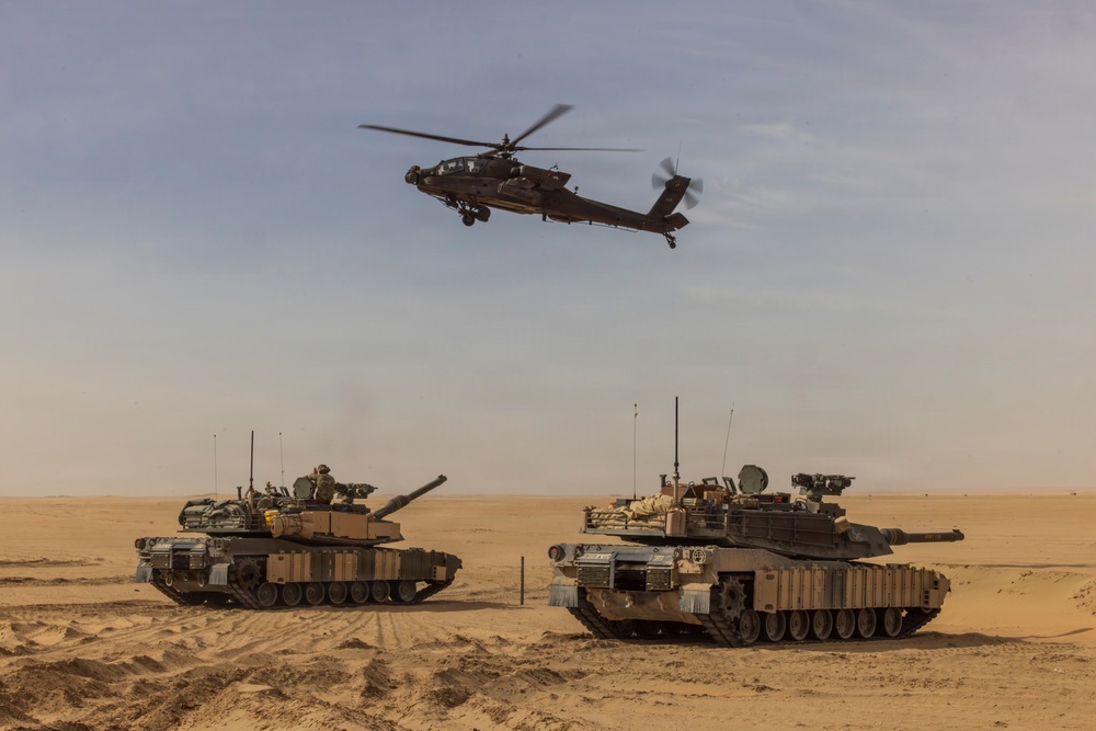 The image shows two main battle tanks positioned on a desert terrain with an attack helicopter flying overhead. The scene depicts a combined arms military operation or exercise in an arid environment. The equipment appears to be in operational condition with crew members visible on at least one of the tanks.