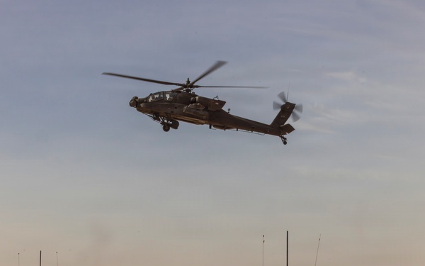 The image shows two main battle tanks positioned on a desert terrain with an attack helicopter flying overhead. The scene depicts a combined arms military operation or exercise in an arid environment. The equipment appears to be in operational condition with crew members visible on at least one of the tanks.