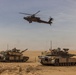 The image shows two main battle tanks positioned on a desert terrain with an attack helicopter flying overhead. The scene depicts a combined arms military operation or exercise in an arid environment. The equipment appears to be in operational condition with crew members visible on at least one of the tanks.