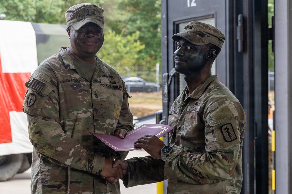 Two U.S. Army soldiers in OCP (Operational Camouflage Pattern) uniforms are shaking hands while jointly holding a purple/maroon certificate folder, suggesting an award or recognition ceremony. The soldier on the left, identified by nametape as 'COLLINS,' appears to be a senior NCO presenting the document. The setting appears to be an outdoor military installation or field environment.