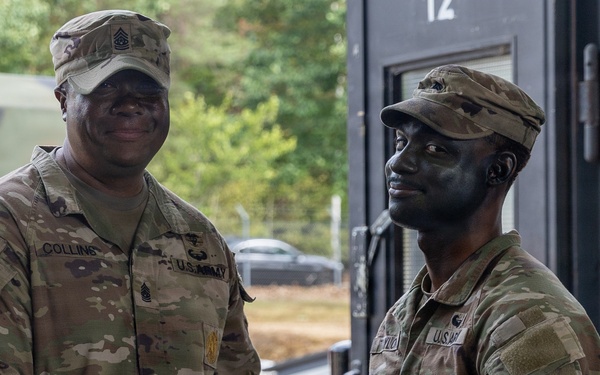 Two U.S. Army soldiers in OCP (Operational Camouflage Pattern) uniforms are shaking hands while jointly holding a purple/maroon certificate folder, suggesting an award or recognition ceremony. The soldier on the left, identified by nametape as 'COLLINS,' appears to be a senior NCO presenting the document. The setting appears to be an outdoor military installation or field environment.