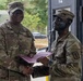Two U.S. Army soldiers in OCP (Operational Camouflage Pattern) uniforms are shaking hands while jointly holding a purple/maroon certificate folder, suggesting an award or recognition ceremony. The soldier on the left, identified by nametape as 'COLLINS,' appears to be a senior NCO presenting the document. The setting appears to be an outdoor military installation or field environment.