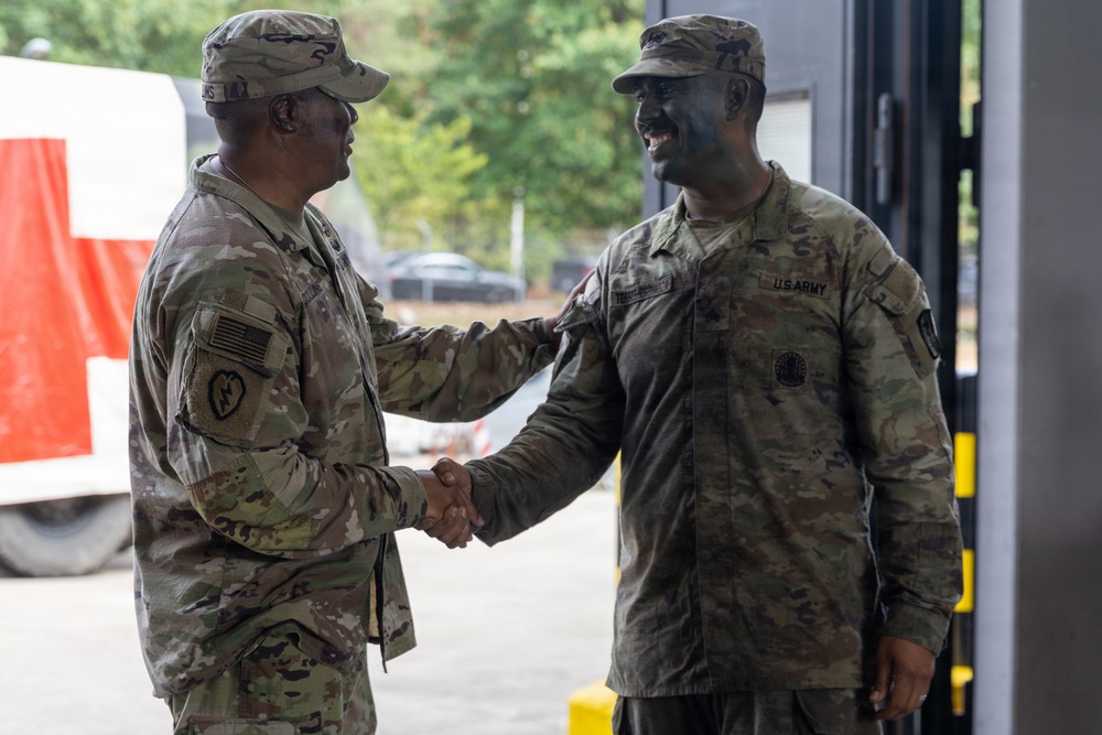 Two U.S. Army soldiers in Operational Camouflage Pattern (OCP) uniforms are shaking hands in what appears to be a military facility or base. The soldier on the right is smiling broadly and has camouflage face paint applied, suggesting recent field training activity. The interaction appears cordial and professional, possibly a greeting or recognition moment.