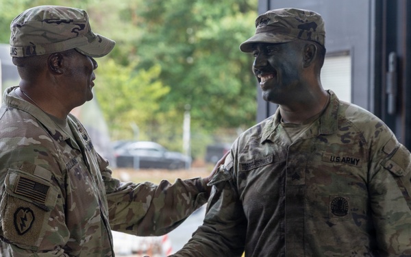 Two U.S. Army soldiers in Operational Camouflage Pattern (OCP) uniforms are shaking hands in what appears to be a military facility or base. The soldier on the right is smiling broadly and has camouflage face paint applied, suggesting recent field training activity. The interaction appears cordial and professional, possibly a greeting or recognition moment.