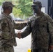 Two U.S. Army soldiers in Operational Camouflage Pattern (OCP) uniforms are shaking hands in what appears to be a military facility or base. The soldier on the right is smiling broadly and has camouflage face paint applied, suggesting recent field training activity. The interaction appears cordial and professional, possibly a greeting or recognition moment.