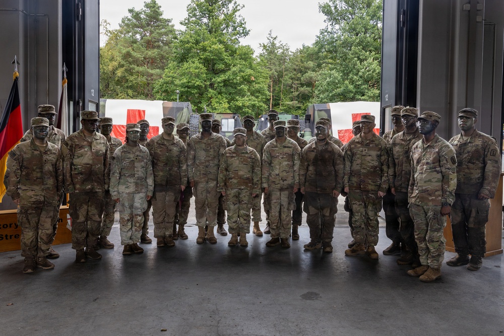A group of approximately 20-25 military personnel in US Army Operational Camouflage Pattern (OCP) uniforms pose for a group photograph inside a large vehicle bay or hangar. The soldiers have face camouflage paint applied, suggesting they are returning from or preparing for a field exercise. A German flag is visible on the left side, indicating this photo was likely taken at a US military installation in Germany.