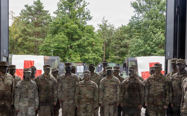 A group of approximately 20-25 military personnel in US Army Operational Camouflage Pattern (OCP) uniforms pose for a group photograph inside a large vehicle bay or hangar. The soldiers have face camouflage paint applied, suggesting they are returning from or preparing for a field exercise. A German flag is visible on the left side, indicating this photo was likely taken at a US military installation in Germany.
