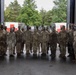 A group of approximately 20-25 military personnel in US Army Operational Camouflage Pattern (OCP) uniforms pose for a group photograph inside a large vehicle bay or hangar. The soldiers have face camouflage paint applied, suggesting they are returning from or preparing for a field exercise. A German flag is visible on the left side, indicating this photo was likely taken at a US military installation in Germany.