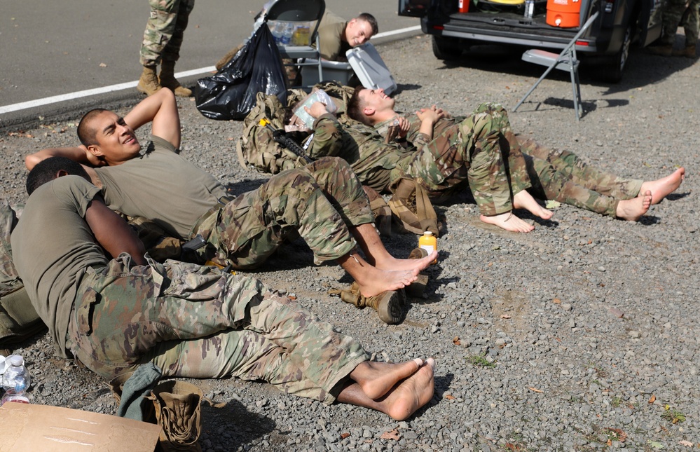 Several soldiers in camouflage uniforms are resting on a gravel surface, with boots and socks removed, suggesting a break after physical exertion. They are lying on their backs or sides near military gear, backpacks, and a vehicle in the background. The scene appears to be a rest stop during a field exercise or training operation.