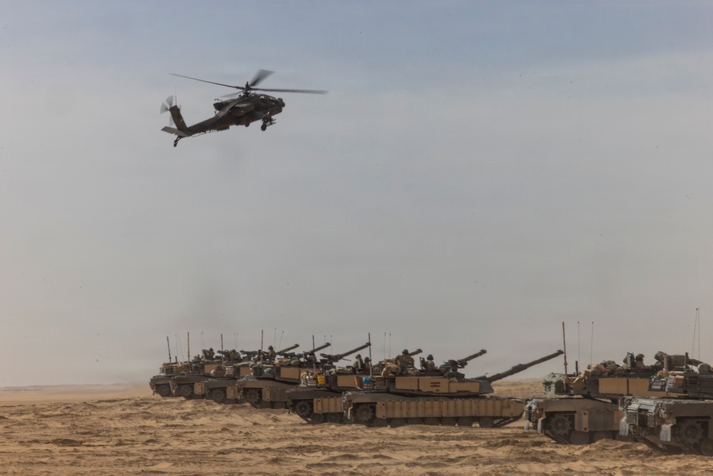 A military scene in a desert environment showing a formation of main battle tanks on the ground with an attack helicopter flying overhead. The tanks appear to be in a staged or operational formation across sandy terrain. The image depicts a combined arms military operation or exercise involving armor and aviation assets.