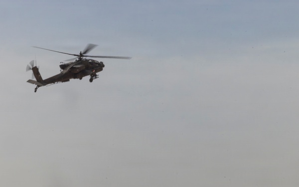 A military scene in a desert environment showing a formation of main battle tanks on the ground with an attack helicopter flying overhead. The tanks appear to be in a staged or operational formation across sandy terrain. The image depicts a combined arms military operation or exercise involving armor and aviation assets.