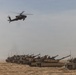A military scene in a desert environment showing a formation of main battle tanks on the ground with an attack helicopter flying overhead. The tanks appear to be in a staged or operational formation across sandy terrain. The image depicts a combined arms military operation or exercise involving armor and aviation assets.