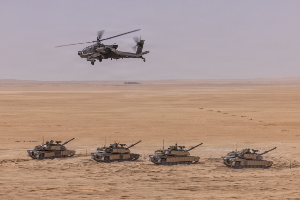 A military exercise or operation is depicted in a desert environment, showing four main battle tanks advancing in a line formation across sandy terrain. An attack helicopter flies low overhead, providing aerial support or coordination. The scene suggests a combined arms operation integrating ground armor and aviation assets.
