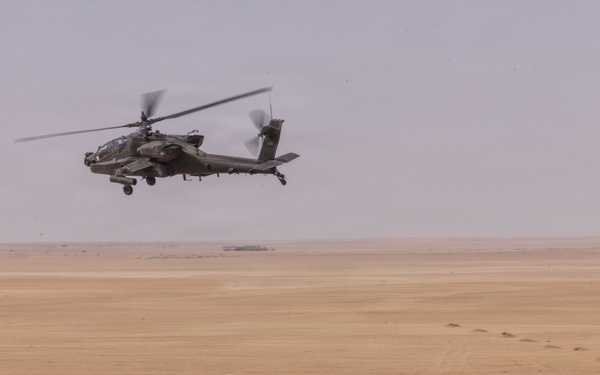 A military exercise or operation is depicted in a desert environment, showing four main battle tanks advancing in a line formation across sandy terrain. An attack helicopter flies low overhead, providing aerial support or coordination. The scene suggests a combined arms operation integrating ground armor and aviation assets.