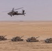 A military exercise or operation is depicted in a desert environment, showing four main battle tanks advancing in a line formation across sandy terrain. An attack helicopter flies low overhead, providing aerial support or coordination. The scene suggests a combined arms operation integrating ground armor and aviation assets.