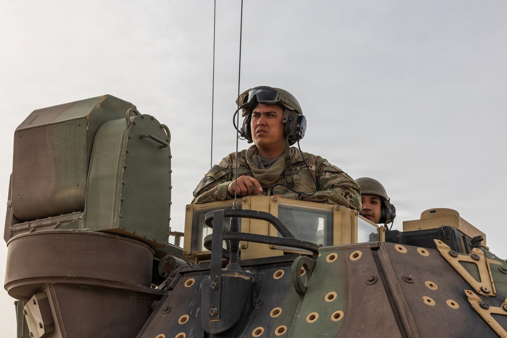 Two U.S. Army soldiers are positioned atop a military armored vehicle, with one soldier standing upright through a hatch wearing a combat helmet with communications headset and OCP (Operational Camouflage Pattern) uniform. A name tape reading 'REEC' or similar and 'U.S. ARMY' branch tape are visible on the primary soldier's uniform. The image is taken from a low angle against an overcast sky, emphasizing the military equipment and personnel.