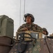 Two U.S. Army soldiers are positioned atop a military armored vehicle, with one soldier standing upright through a hatch wearing a combat helmet with communications headset and OCP (Operational Camouflage Pattern) uniform. A name tape reading 'REEC' or similar and 'U.S. ARMY' branch tape are visible on the primary soldier's uniform. The image is taken from a low angle against an overcast sky, emphasizing the military equipment and personnel.