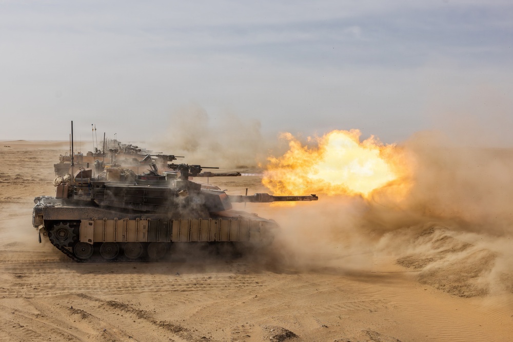 A column of main battle tanks is operating in a desert environment, with the lead tank actively firing its main gun. A large muzzle blast with bright orange flame and smoke is visible at the end of the barrel. Multiple tanks are visible in formation extending into the background.