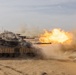 A column of main battle tanks is operating in a desert environment, with the lead tank actively firing its main gun. A large muzzle blast with bright orange flame and smoke is visible at the end of the barrel. Multiple tanks are visible in formation extending into the background.