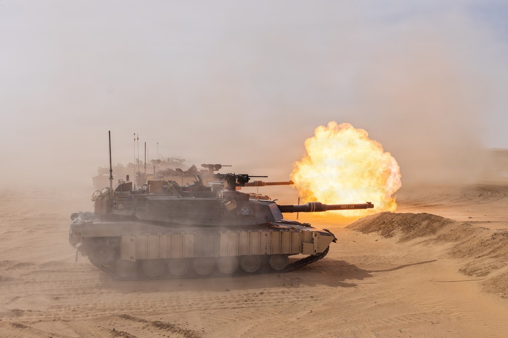 A main battle tank is captured mid-fire in a desert environment, with a large muzzle blast and fireball visible at the end of the gun barrel. The scene shows dusty, sandy terrain with at least one additional armored vehicle visible in the background. The image appears to document a live-fire military exercise or combat operation.