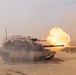 A main battle tank is captured mid-fire in a desert environment, with a large muzzle blast and fireball visible at the end of the gun barrel. The scene shows dusty, sandy terrain with at least one additional armored vehicle visible in the background. The image appears to document a live-fire military exercise or combat operation.