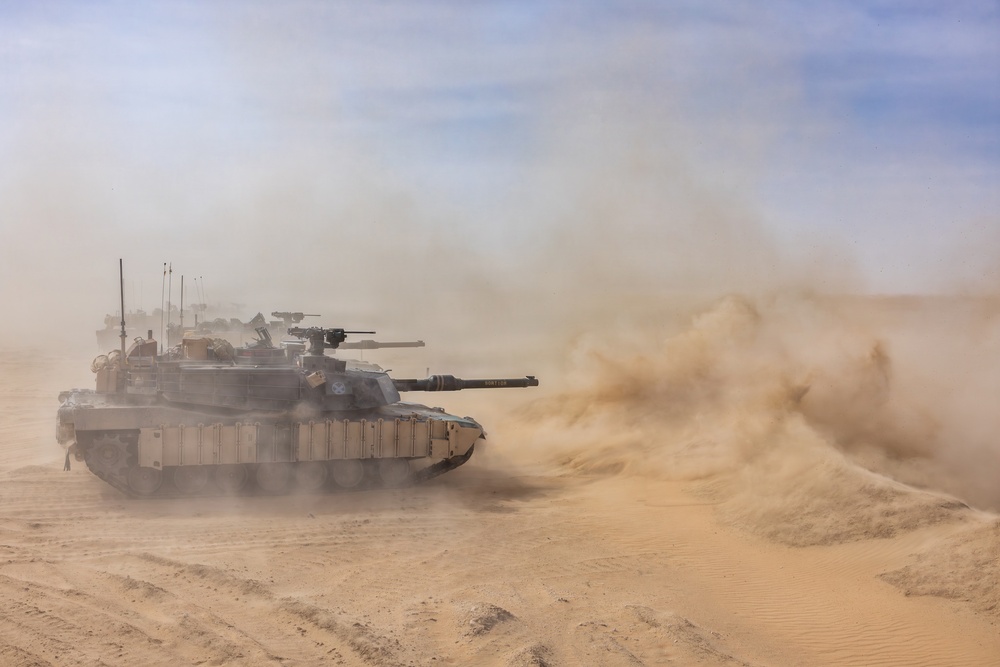 A main battle tank, consistent with an M1 Abrams, is maneuvering at speed across a desert terrain, generating large dust clouds from its tracks. At least one additional armored vehicle is visible in the background through the dust haze. The scene appears to depict a military training exercise or operational movement in an arid environment.