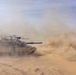 A main battle tank, consistent with an M1 Abrams, is maneuvering at speed across a desert terrain, generating large dust clouds from its tracks. At least one additional armored vehicle is visible in the background through the dust haze. The scene appears to depict a military training exercise or operational movement in an arid environment.