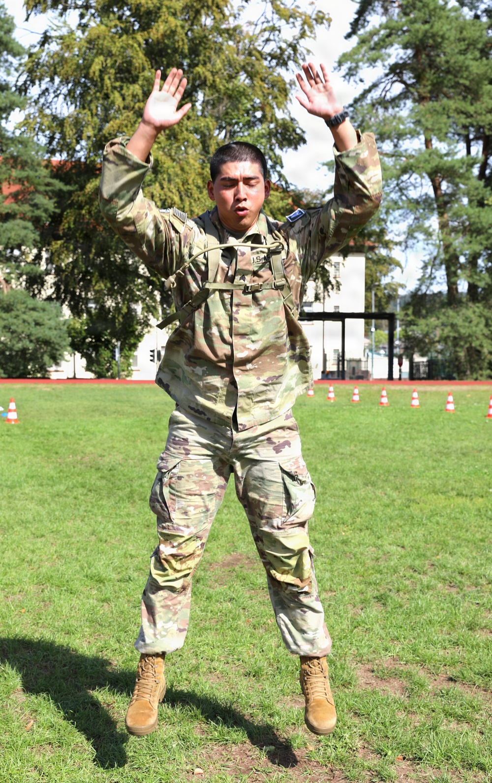 A U.S. Army soldier in Operational Camouflage Pattern (OCP) uniform is captured mid-jump performing what appears to be a jumping jack or physical training exercise on a grass field. The soldier is wearing a rucksack/backpack and tan combat boots, with both arms raised overhead and legs spread apart. Orange traffic cones are visible in the background, suggesting an organized physical fitness or training event.