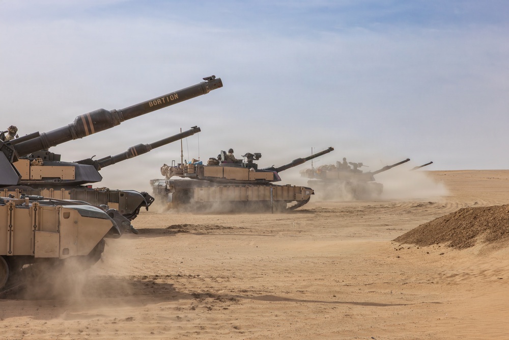A column of M1 Abrams main battle tanks is moving through a desert environment, kicking up dust as they advance. The tanks are painted in desert tan camouflage and appear to be conducting a military exercise or operation. At least four tanks are visible, with the lead tank bearing the name 'BORTION' stenciled on its gun barrel.