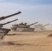 A column of M1 Abrams main battle tanks is moving through a desert environment, kicking up dust as they advance. The tanks are painted in desert tan camouflage and appear to be conducting a military exercise or operation. At least four tanks are visible, with the lead tank bearing the name 'BORTION' stenciled on its gun barrel.