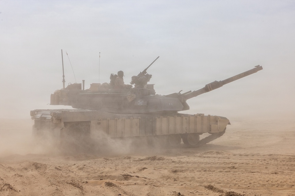 A main battle tank is moving through a desert environment, kicking up significant dust and sand. At least one crew member is visible in the open hatch on top of the turret. The tank appears to be in desert camouflage coloring and is equipped with side skirt armor panels.