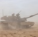 A main battle tank is moving through a desert environment, kicking up significant dust and sand. At least one crew member is visible in the open hatch on top of the turret. The tank appears to be in desert camouflage coloring and is equipped with side skirt armor panels.