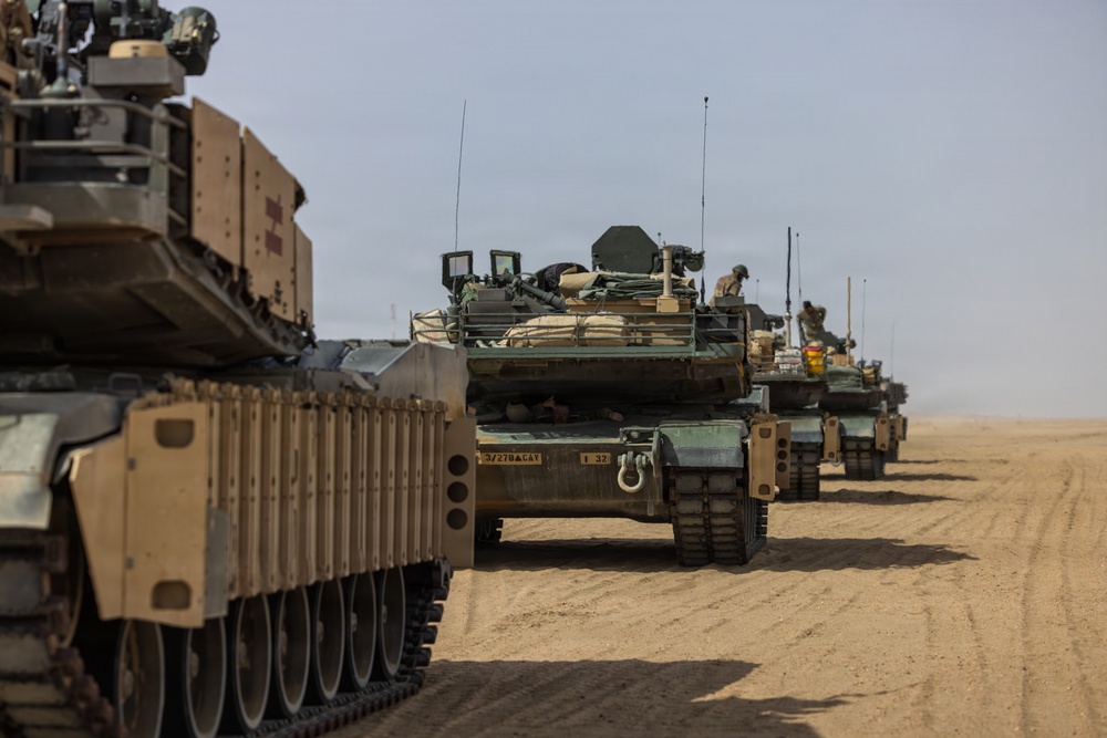A column of M1 Abrams main battle tanks is lined up on a desert terrain, moving or halted in formation. The tanks are fitted with desert-tan paint schemes and additional equipment including reactive armor panels and external gear. At least one unit marking is visible on the second tank reading '3/270 ACAV' and 'I 32'.