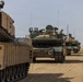 A column of M1 Abrams main battle tanks is lined up on a desert terrain, moving or halted in formation. The tanks are fitted with desert-tan paint schemes and additional equipment including reactive armor panels and external gear. At least one unit marking is visible on the second tank reading '3/270 ACAV' and 'I 32'.