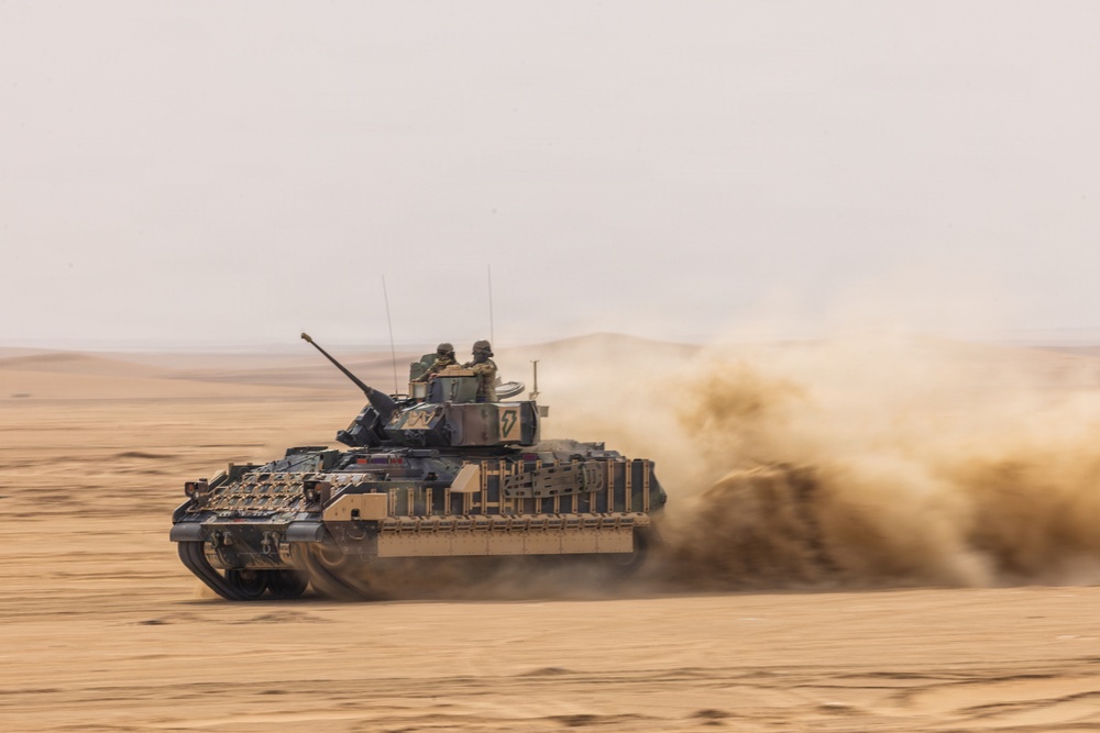 A tracked armored fighting vehicle is moving at speed across a flat desert terrain, kicking up a large dust cloud behind it. Two crew members are visible in the open hatches of the turret. The vehicle appears to be in an active operational or training environment based on its movement and desert setting.