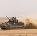 A tracked armored fighting vehicle is moving at speed across a flat desert terrain, kicking up a large dust cloud behind it. Two crew members are visible in the open hatches of the turret. The vehicle appears to be in an active operational or training environment based on its movement and desert setting.