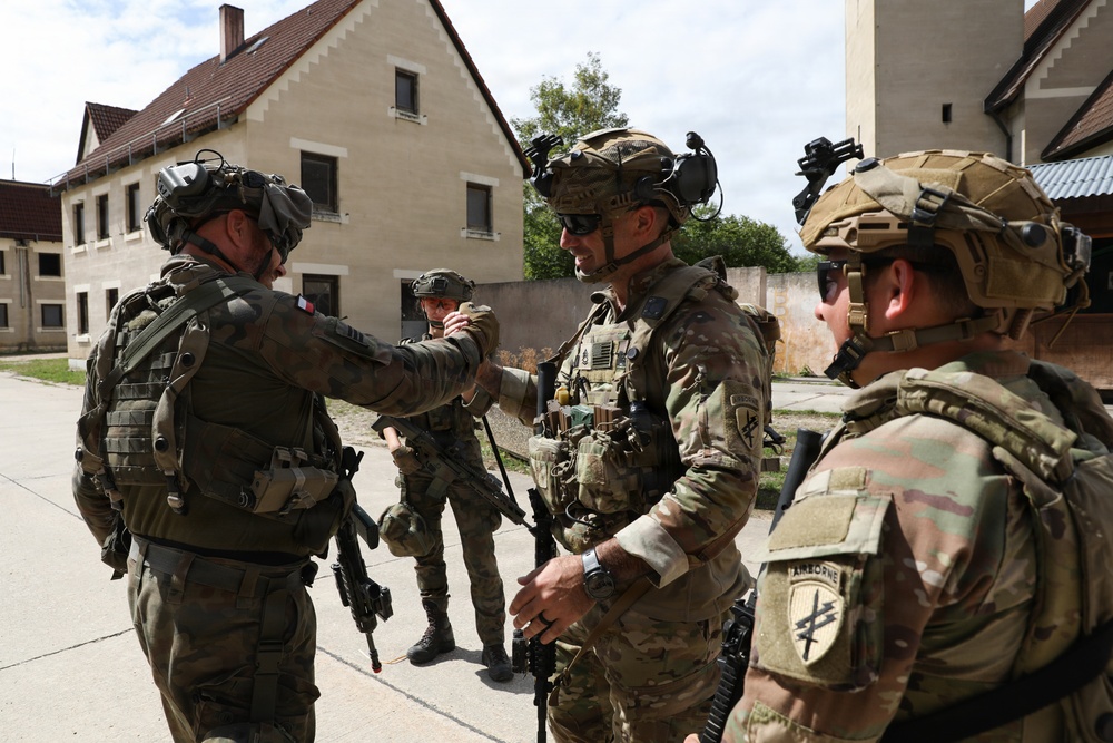 The image shows a group of military personnel in full combat gear interacting in what appears to be a training or operational environment near European-style buildings. Soldiers wearing different national insignia, including what appears to be a French flag patch and U.S. Army Airborne patches, are engaged in a briefing or coordination exchange. The scene suggests a multinational military exercise or joint operation.